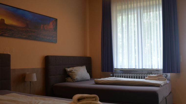 A simple room with two beds and a window that lets in a lot of natural light. A picture of a landscape hangs on the wall.