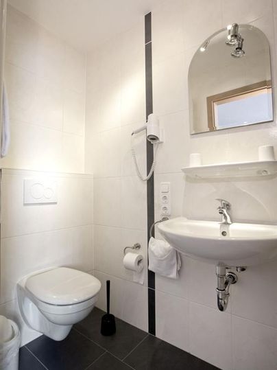 A modern bathroom with white tiles and a wall-mounted sink. There is a toilet and a handy shelf for toiletries.
