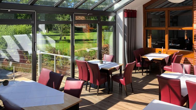Un restaurant lumineux avec de grandes fenêtres et des chaises rouges. Les tables sont dressées et la vue donne sur la verdure.