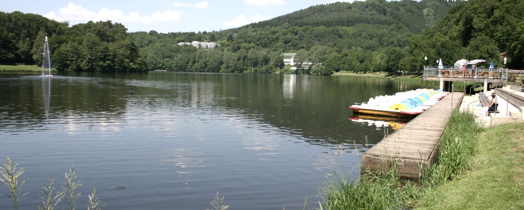 Le lac de barrage de Bitburg aux eaux calmes, entour&eacute; de collines verdoyantes. Des p&eacute;dalos sont amarr&eacute;s au ponton, des gens se d&eacute;tendent sur la rive. Une belle journ&eacute;e d'&eacute;t&eacute;., &copy; TI Bitburger Land