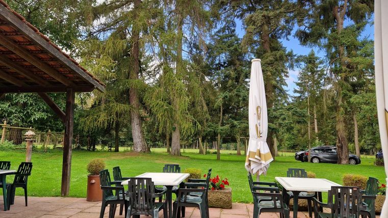 Ein einladender Außenbereich mit Tischen und Stühlen in einer grünen Umgebung. Im Hintergrund sind hohe Bäume und ein Auto sichtbar.