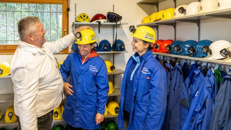 Drei Personen stehen in einem Raum und tragen Arbeitskleidung sowie Helme. Sie scheinen sich auf eine Sicherheitsunterweisung oder ein Training vorzubereiten.