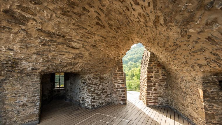Ein Innenraum mit einer gewölbten Steinmauer und einem Holzfußboden. Durch die Fenster sieht man die umgebende Landschaft.